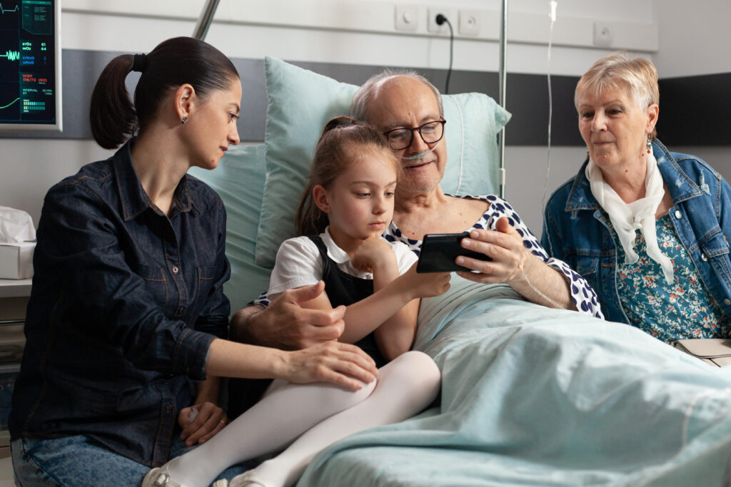 Grandfather browsing on internet with little granddaughter using modern smartphone in hospital ward during clinical therapy. Sick elderly senior man patient resting in bed, healthcare support